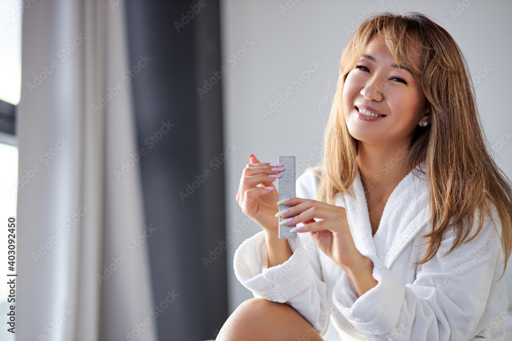woman files up her nails while sitting on the bed wearing white bathrobe, after shower, at home, relax time