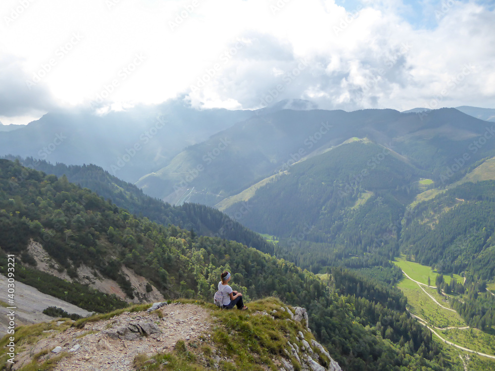 A backpacker woman sitting at the mountain ledge with the panoramic view on Austrian Alps. Mountains overgrown with forest. High and sharp mountains are shrouded with thick fog. Outdoor activities