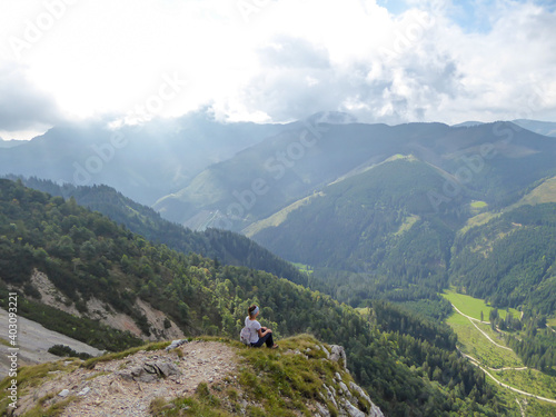 A backpacker woman sitting at the mountain ledge with the panoramic view on Austrian Alps. Mountains overgrown with forest. High and sharp mountains are shrouded with thick fog. Outdoor activities