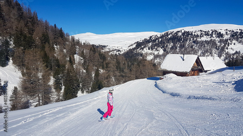 A snowboarding woman in pink outfit going down the slope in Innerkrems, Austria. There is s snow-capped cottage along the ski run. Dense forest on the side. High mountains in the back. Winter outdoor photo