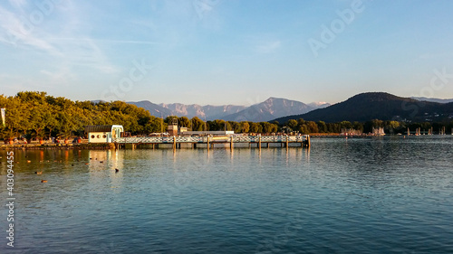A panoramic capture of the pier on the Woerthersee lake in Austria. In the back there are high Alpine chains. The shore of the lake is overgrown with dense forest. Serenity and calmness. Golden hour. © Chris