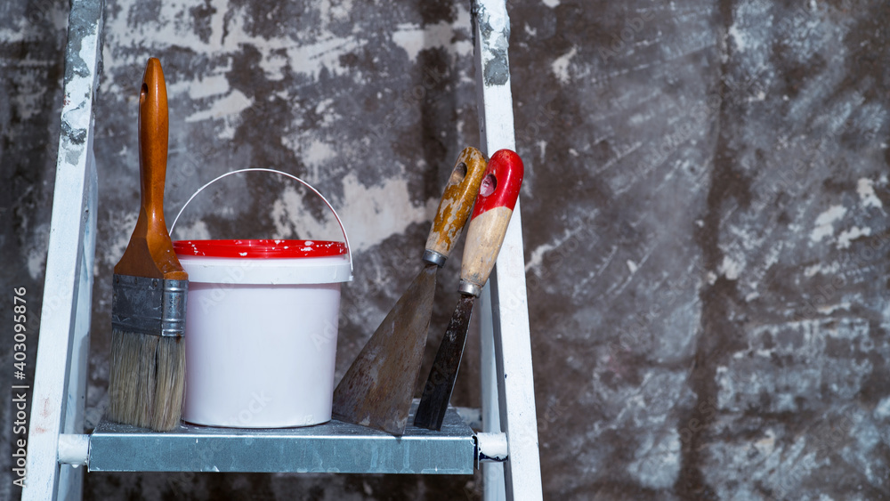 A brush, a bucket with a red lid and spatulas stand on a metal ladder. Background. A brush, a bucket with a red lid and spatulas stand on a metal ladder. Background.