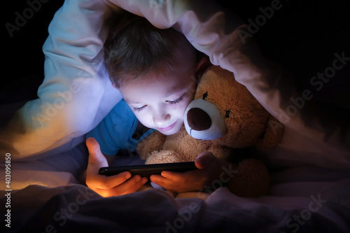 happy boy lies with toy bear in bed under a blanket and using a digital tablet smartphone device in the dark.