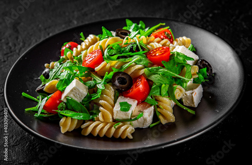 Salad with pasta, arugula, tomato, cheese and olives on black plate on Dark grey black slate background