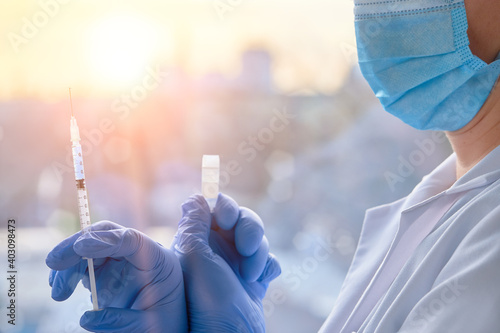 Medic, doctor or nurse with covid-19 vaccine vial and syringe. Mature doctor in face mask, glasses, hat, blue gloves and protective white gown. Romantic view over town with sunset on background.