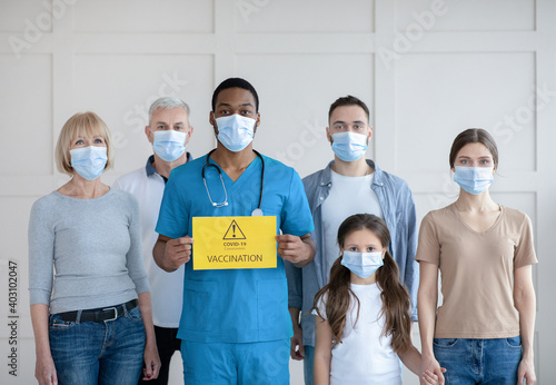 Portrait of diverse people in face masks standing behind black doctor with Covid-19 vaccination sign at clinic