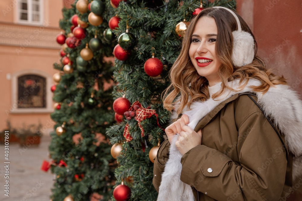 Lady in a knitted sweater and fluffy headphones. Outdoor portrait of young beautiful happy smiling girl is wearing fur coat near decorated Christmas tree. Lady in a knitted sweater and fluffy headphones. Outdoor portrait of young beautiful happy smiling girl is wearing fur coat near decorated Christmas tree.