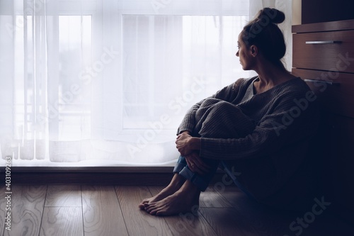 Sad woman sitting on the floor in her living room looking out the window. Autumn depression. © Daniel Jędzura