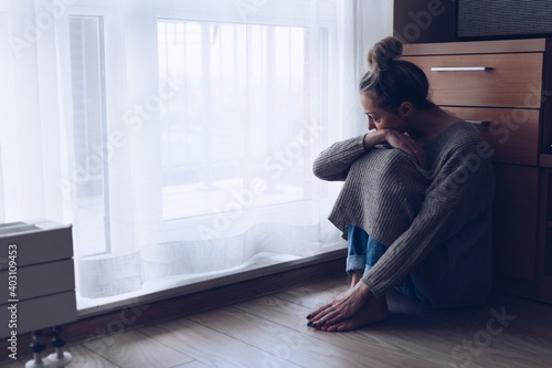 Obraz na płótnie A devastated sad woman is sitting on the floor in her living room looking at the window. She is depressed. w salonie