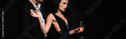 brunette woman in dress holding glass of champagne and pulling away man in suit isolated on black, banner