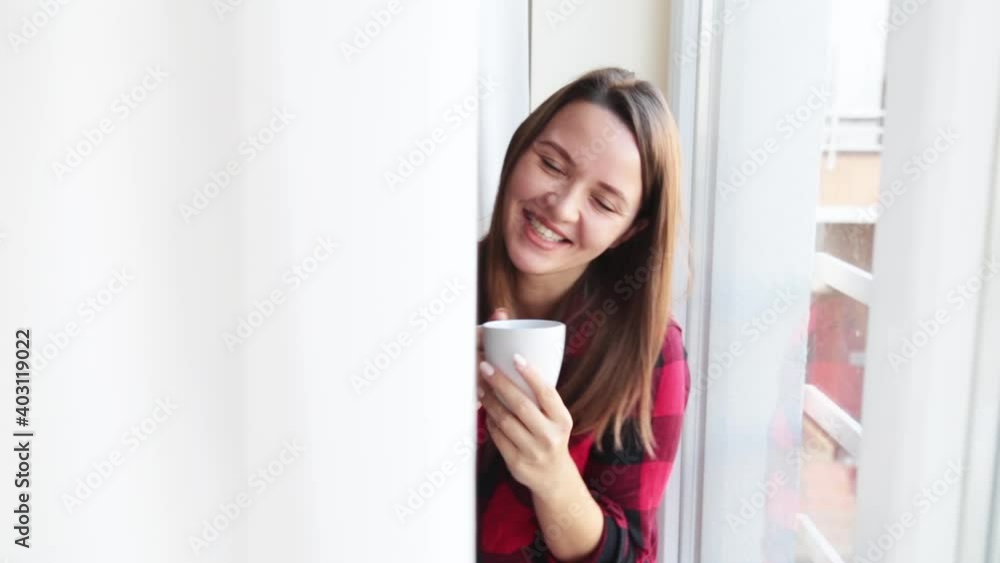 portrait of young cheerful girl sitting on the windowsill next to the window with cup of tea. Young woman drinking hot coffee looking at camera and laughing . stay home happiness concept portrait of young cheerful girl sitting on the windowsill next to the window with cup of tea. Young woman drinking hot coffee looking at camera and laughing . stay home happiness concept