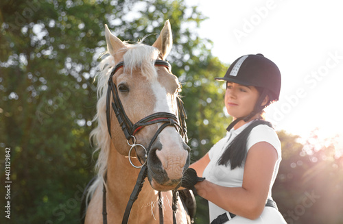 Fototapeta Naklejka Na Ścianę i Meble -  Young woman in horse riding suit and her beautiful pet outdoors on sunny day