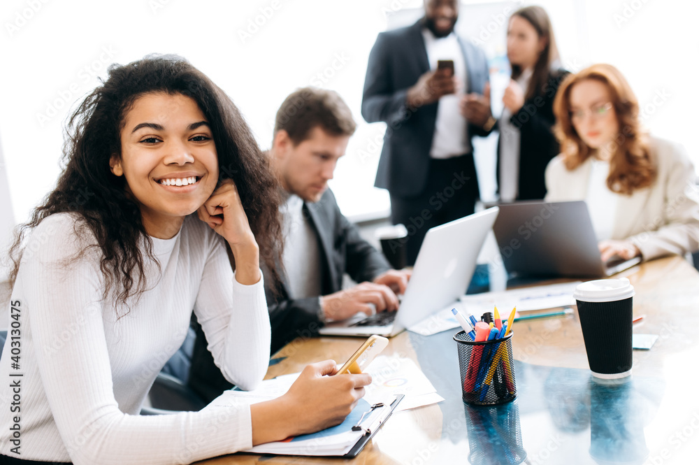 Young african american business woman at the workplace. Beautiful female employee sit at the work desk in office, hold a smartphone in hand. Smiling woman at work briefing meeting Young african american business woman at the workplace. Beautiful female employee sit at the work desk in office, hold a smartphone in hand. Smiling woman at work briefing meeting