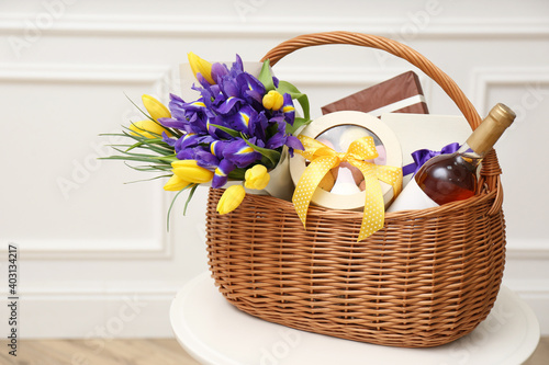 Wicker basket with gifts, bouquet and wine on white table indoors photo