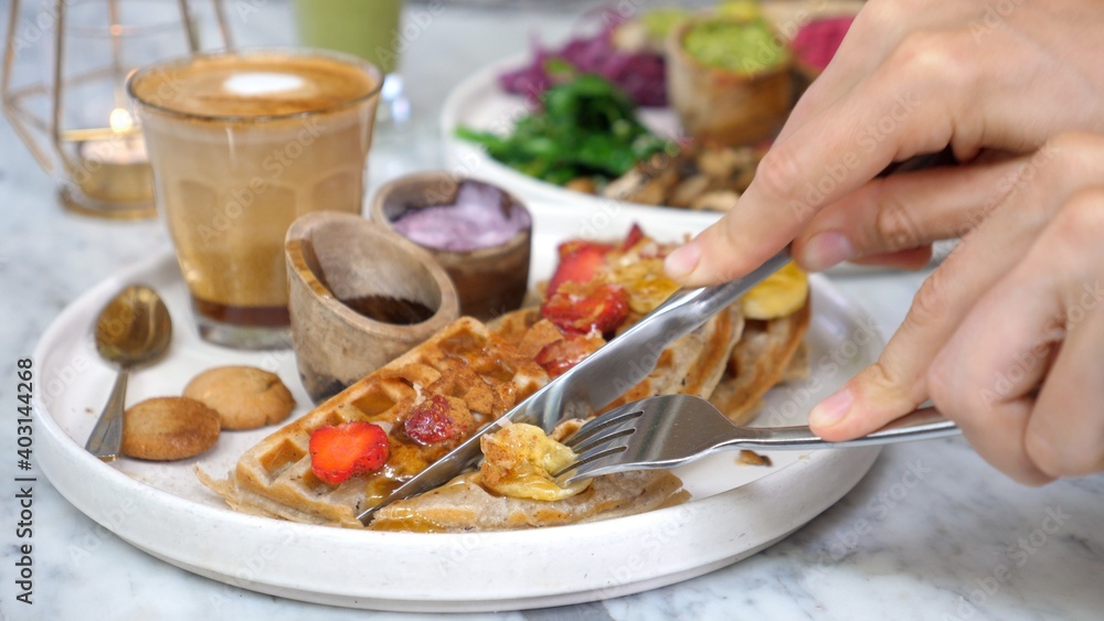 Closeup of the female's hands cutting vegan waffles on the marble table served with vegan dish and latte coffee.