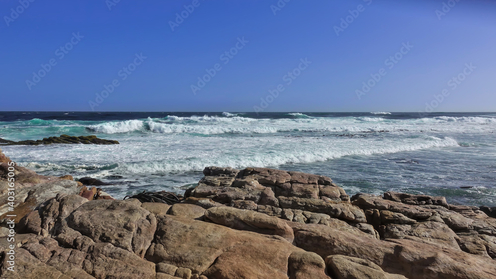 The turquoise waves of the Atlantic Ocean leave a white foam on the coast. Ancient boulders in the foreground. Clear blue sky. Cape of Good Hope. South Africa