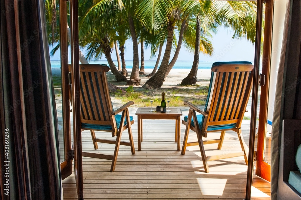 Beach view with palm trees and ocean from the hotel villa window. Wooden chairs with table and champagne
