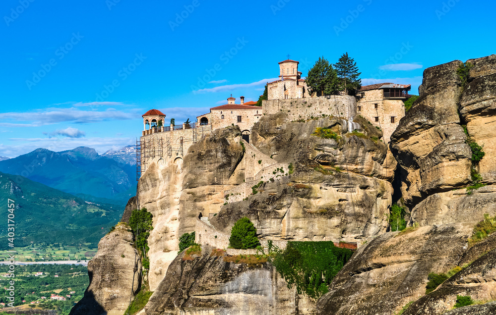 Close view of clifftop Varlaam monastery and rocky slopes of Meteora valley lit by morning sun. Village and mountains. Greece, UNESCO World Heritage