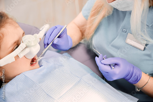 Female dentist checking child teeth with dental explorer and mirror while girl lying in dental chair with inhalation sedation. Concept of pediatric, sedation dentistry and dental care. photo