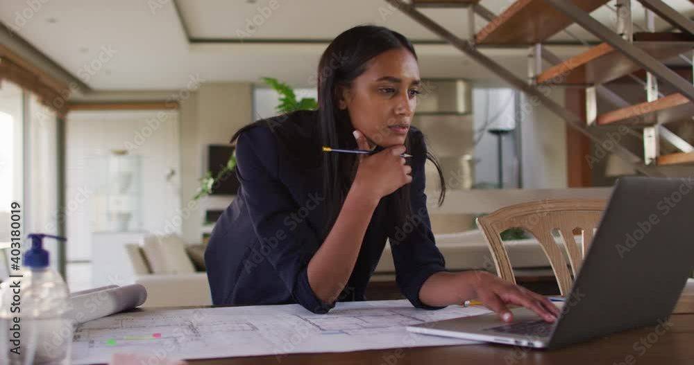 Mixed race woman by desk at home using laptop looking through blueprints Mixed race woman by desk at home using laptop looking through blueprints