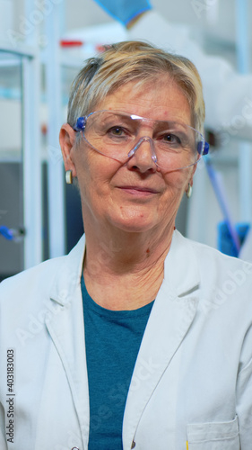 Portrait of old scientist woman smiling at camera in modern equipped lab. Multiethnic team examining virus evolution using high tech and chemistry tools for scientific research, vaccine development.