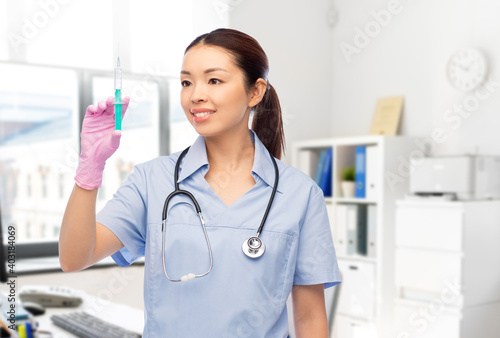 medicine, vaccination and healthcare concept - happy smiling asian female doctor or nurse in blue uniform with stethoscope and syringe over hospital background