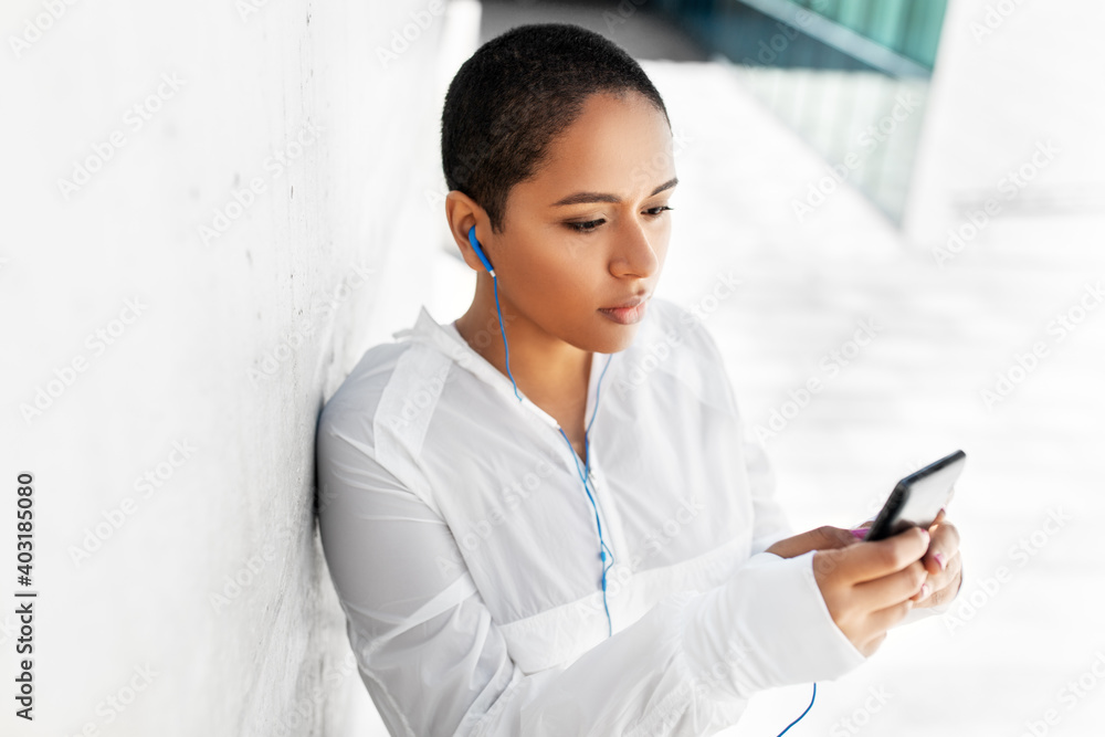 fitness, sport and technology concept - young african american woman with earphones listening to music on smartphone outdoors