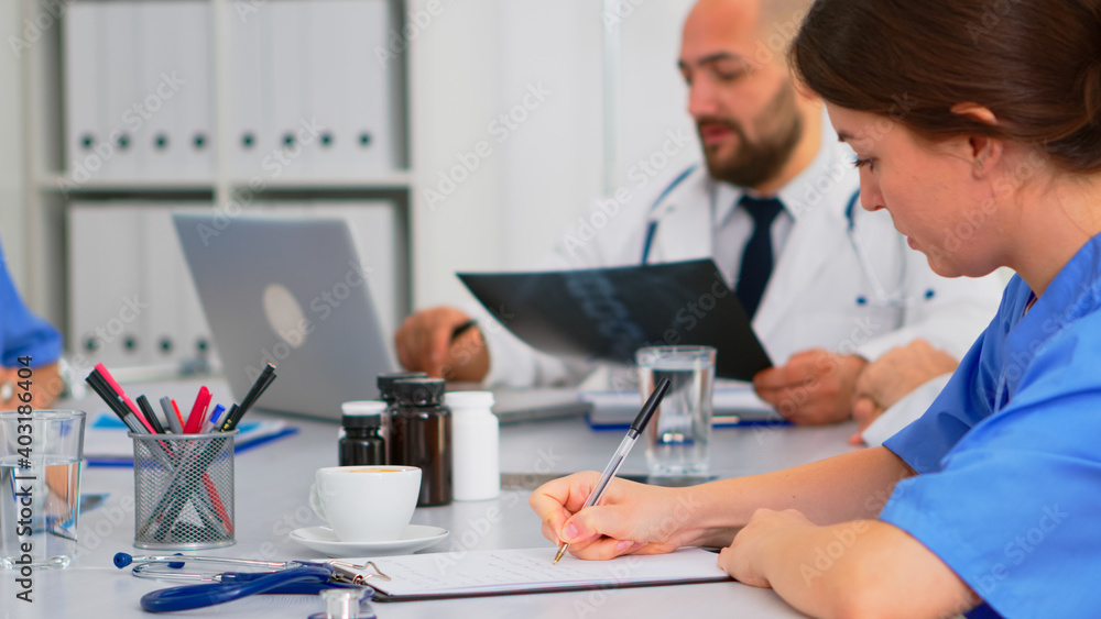 Close up of nurse taking notes on clipboard while radiologist coworkers discussing in background analysing x-ray and writing on laptop. Profesional teamworkers having medical meeting, brainstorming