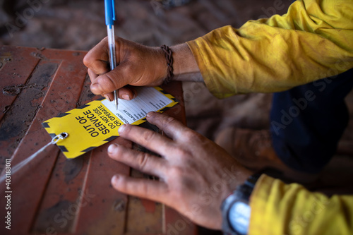 Safe workplaces practices construction worker wearing long sleeve shirt using a blue pen writing on yellow out of service tag prior tagging on damaged faulty goods equipment     