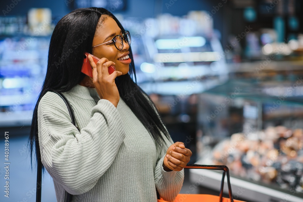 Fototapeta premium African american woman with shopping cart trolley in the supermarket store speak on mobile phone.