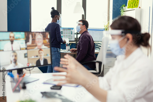 African woman having a conversation with her coworker waring face mask at workplace wearing face mask. Entrepreneur having videocall while colleagues working respecting social distance during global