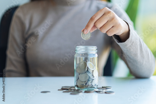 Closeup image of a hand collecting and putting coins in a glass jar for saving money concept