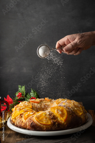 View of woman's hand pouring sugar on Roscon de Reyes, homemade, on dark background, in portrait, with copy space photo