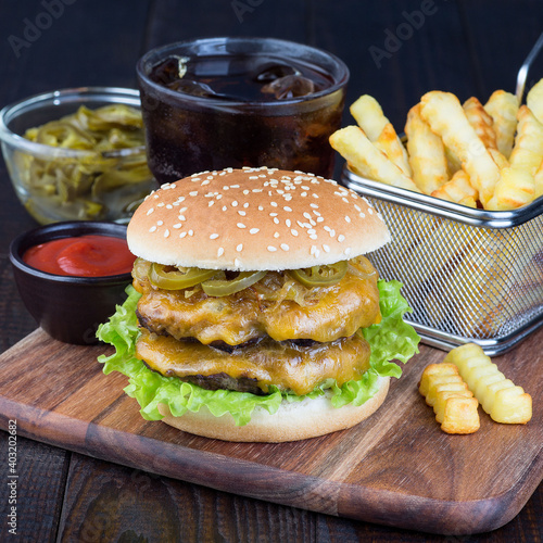 Double cheese burger with caramelized onion, lettuce and jalapeno, served with french fries and soda, on a wooden board, square format