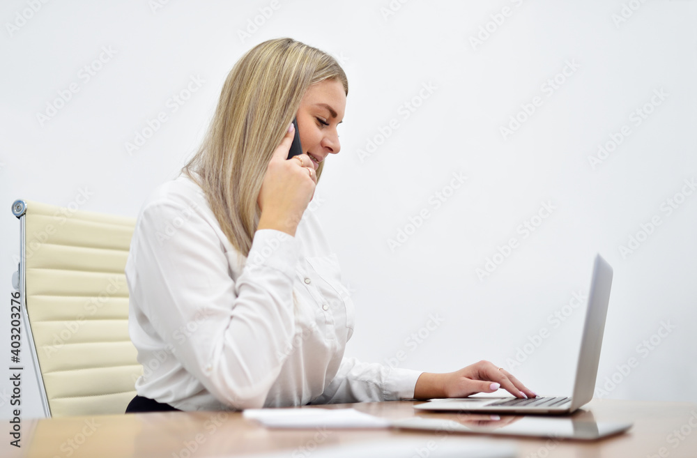 Young caucasian white woman talking on smartphone and working with laptop in office Young caucasian white woman talking on smartphone and working with laptop in office