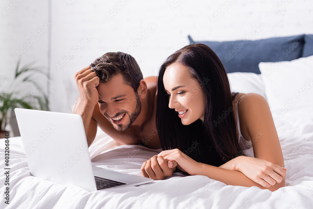 happy young couple laughing while lying on bed near laptop on blurred background happy young couple laughing while lying on bed near laptop on blurred background