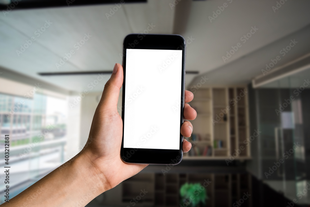 Hand holding white mobile phone with blank white screen in office.