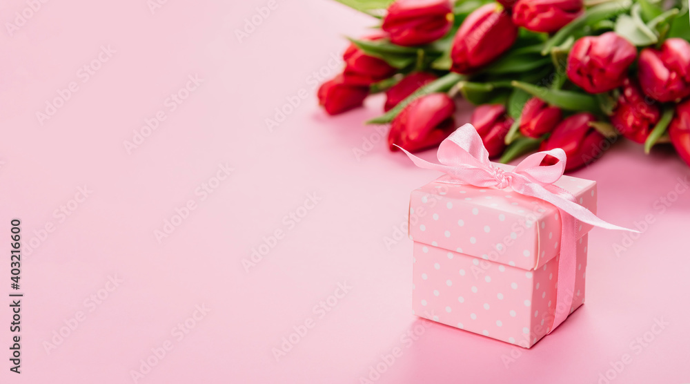 Gift box with a pink ribbon in white polka dots on a background of a bouquet of red tulips with copy space for text. Wallpaper or banner for gift shop, flower shop or jewelry store. Valentine day