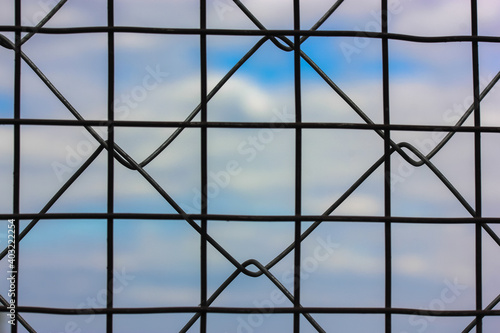 Metal protective grid or netting against a background of blue sky with white clouds. Protected facility  parking lot or prison. High quality photo