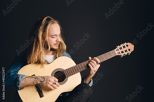 Portrait of long haired tattooed male musician playing on acoustic guitar over black background. Stylish guy with guitar posing in studio