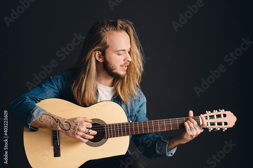 Side view of long haired tattooed guy playing on acoustic guitar and singing preparing for concert isolated on black background. Stylish singer with guitar