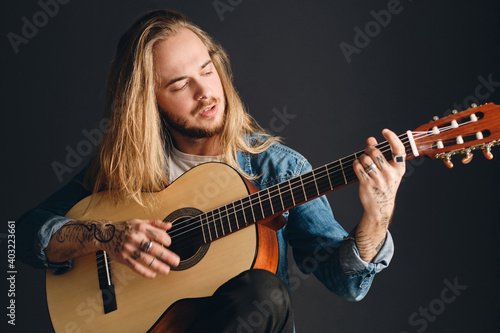 Attractive long haired tattooed musician playing on acoustic guitar preparing for concert over black background