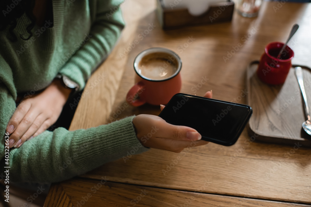 partial view of african american woman holding smartphone with blank screen near cup of cappuccino