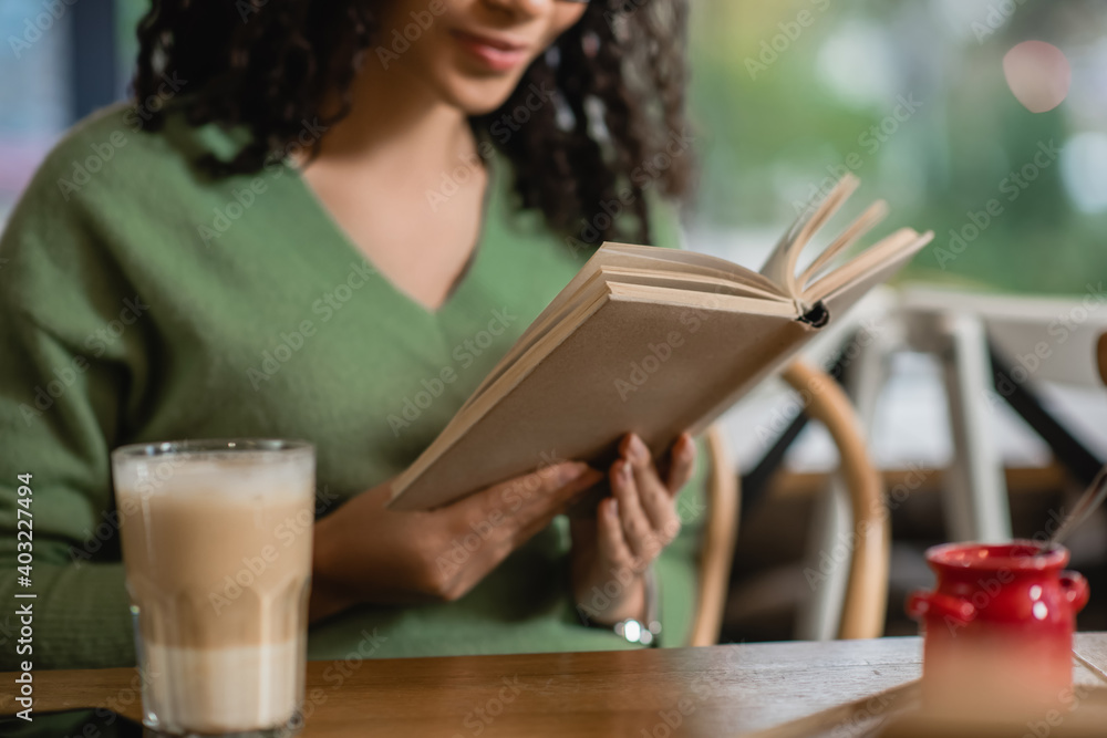 partial view of african american woman reading book near glass with latte on blurred foreground - obrazy, fototapety, plakaty 