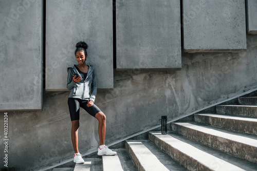 Uses phone. Young african american woman in sportive clothes have workout outdoors at daytime