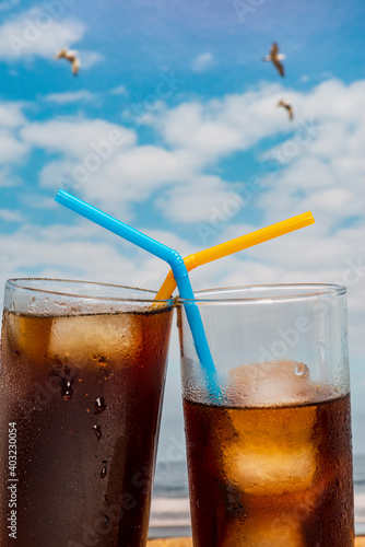 Close-up of two glasses of soda with ice and straws on the beach with blue sky and seagulls flying in Tenerife, Canary Islands.