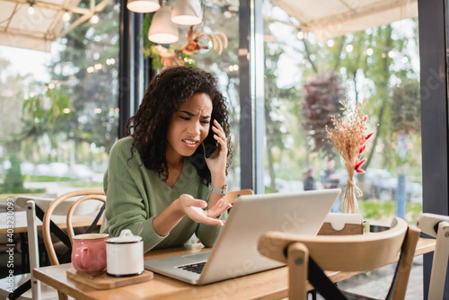 displeased african american woman talking on smartphone and pointing with hand at laptop in cafe