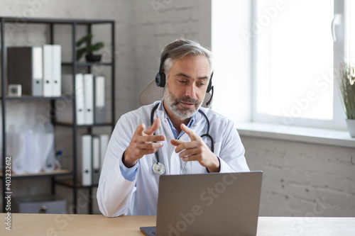 Portrait of senior grey-haired male doctor in his office using laptop for video chat with a patient. Online consultation with doctor for diagnoses and treatment recommendation. Telehealth concept. photo