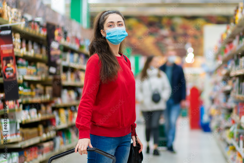 Shopping. Portrait of a young woman in a medical mask poses in the aisle of a supermarket. The concept of consumerism and the new reality