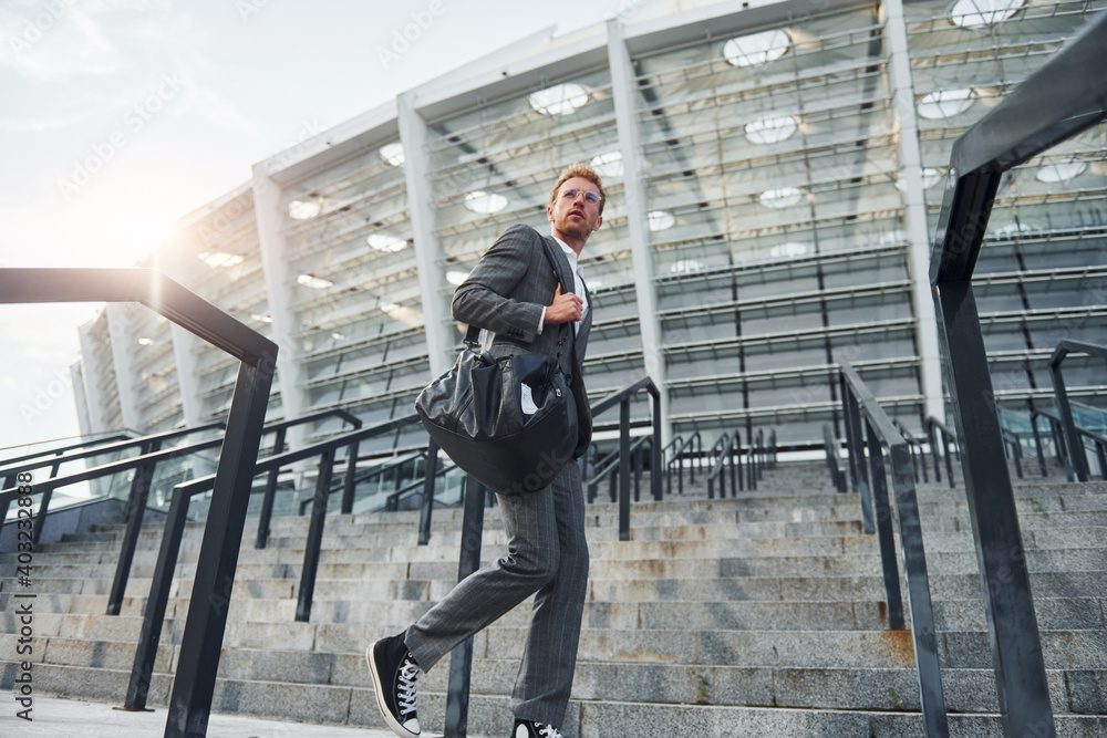 Holds bag in hands. Young businessman in grey formal wear is outdoors in the city Holds bag in hands. Young businessman in grey formal wear is outdoors in the city